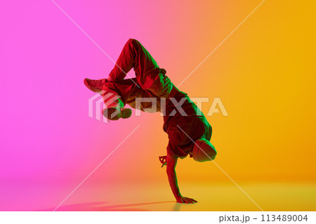 Dynamic photo of young, talented man dancing breakdance moves in neon light against gradient pink-yellow background. 113489004