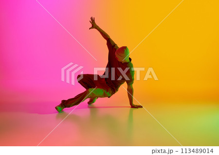 Young man, dressed streetwear performing freestyle moves in motion in neon light against gradient pink-yellow background. 113489014