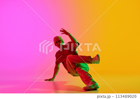 Young athlete man, dressed urban style outfit dancing in motion in neon light against gradient pink-yellow background. Young athlete man, dressed urban style outfit dancing in motion in neon light against gradient pink-yellow background. 113489020