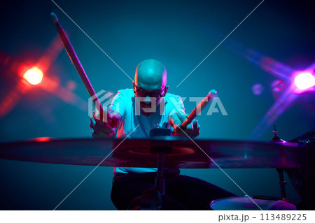 Emotional bald drummer in casual outfit with drumsticks in pink-purple stage lighting against gradient studio background. Emotional bald drummer in casual outfit with drumsticks in pink-purple stage lighting against gradient studio background. 113489225