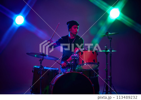 Contemporary drummer playing with joy in pink-purple stage lighting against gradient studio background. 113489232