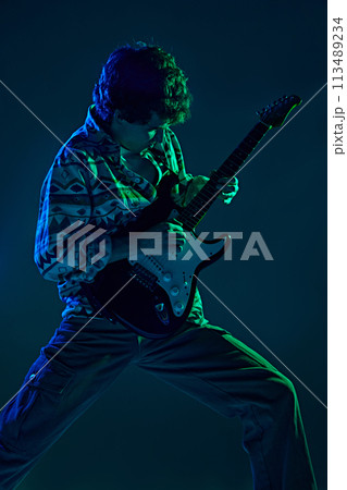 Young talented guitarist in retro attire performing in green blue stage neon light against gradient studio background. Young talented guitarist in retro attire performing in green blue stage neon light against gradient studio background. 113489234