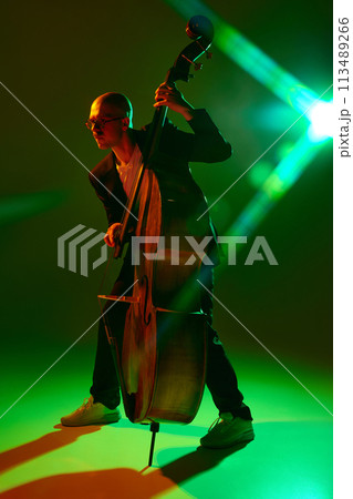 Double bass player performing contemporary jazz composition in red-green neon light against gradient studio background. Double bass player performing contemporary jazz composition in red-green neon light against gradient studio background. 113489266