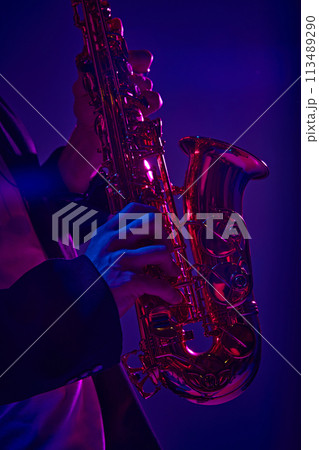 Cropped photo of talented solo player performing on saxophone in vibrant pink neon light against dark studio background. Cropped photo of talented solo player performing on saxophone in vibrant pink neon light against dark studio background. 113489290