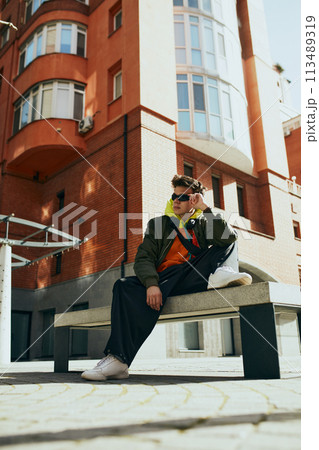 Creative portrait of young attractive man dressed oversize clothes, stylish accessories against cityscape background. 113489319