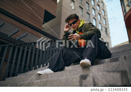 In heart of city's rhythm. Cool attitude young man sitting using phone on stairs against skyline, urban background. In heart of city's rhythm. Cool attitude young man sitting using phone on stairs against skyline, urban background. 113489335