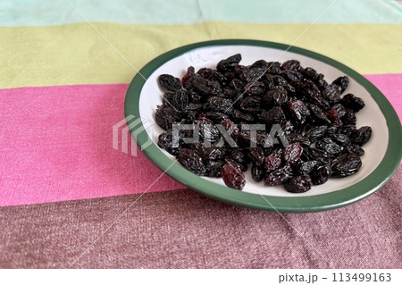 A lot of black raisin in the white green plate is located on the table at home. Close-up A lot of black raisin in the white green plate is located on the table at home. Close-up 113499163
