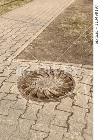 Iron manhole covered dirt above an observation well is located next to pedestrian road during the daytime. Close-up Iron manhole covered dirt above an observation well is located next to pedestrian road during the daytime. Close-up 113499197