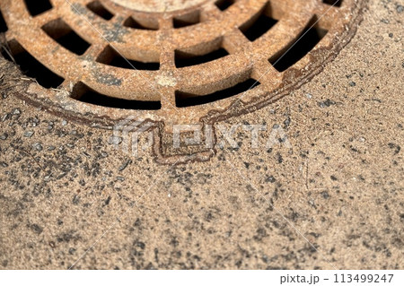 A rusty iron hatch with a pattern above an observation well is located on auto road in daytime. Close-up A rusty iron hatch with a pattern above an observation well is located on auto road in daytime. Close-up 113499247