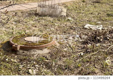 A rusty iron hatch with a pattern covered moss above an observation well is located next to a dry grass in the daytime. Close-up 113499264