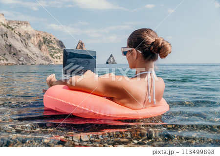 Woman freelancer works on laptop swimming in sea on pink inflatable ring. Happy tourist in sunglasses floating on inflatable donut and working on laptop computer in calm ocean. Remote working anywhere Woman freelancer works on laptop swimming in sea on pink inflatable ring. Happy tourist in sunglasses floating on inflatable donut and working on laptop computer in calm ocean. Remote working anywhere 113499898