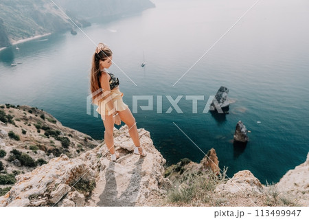Woman travel sea. Happy tourist enjoy taking picture outdoors for memories. Woman traveler looks at the edge of the cliff on the sea bay of mountains, sharing travel adventure journey 113499947