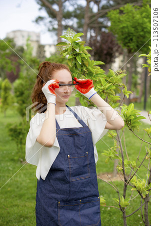 Side view of female florist standing, putting on glasses in garden. Concept of taking care of nature. Side view of female florist standing, putting on glasses in garden. Concept of taking care of nature. 113507106