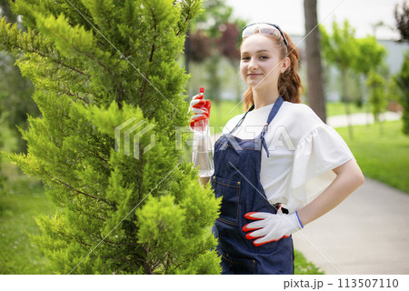 Front view of girl with red hair spraying trees, holding hand on waist. Concept of taking care of plants. Front view of girl with red hair spraying trees, holding hand on waist. Concept of taking care of plants. 113507110