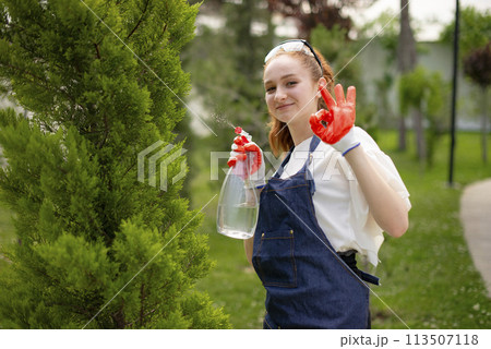 Front view of young female florist standing, spraying trees, showing alright, smiling. Concept of taking care of plants. Front view of young female florist standing, spraying trees, showing alright, smiling. Concept of taking care of plants. 113507118