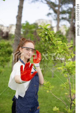 Front view of female florist standing in garden, showing hand, looking at camera. Concept of taking care of nature. Front view of female florist standing in garden, showing hand, looking at camera. Concept of taking care of nature. 113507164