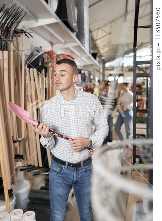 Buying shovel in supermarket - man with interest chooses shovel in hardware store for working in garden Buying shovel in supermarket - man with interest chooses shovel in hardware store for working in garden 113507560