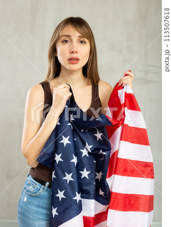 Sad young woman holding flag of the USA against unicoloured background Sad young woman holding flag of the USA against unicoloured background 113507618