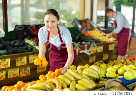 Woman selelr of vegetable department store is replenishing showcase with oranges 113507855