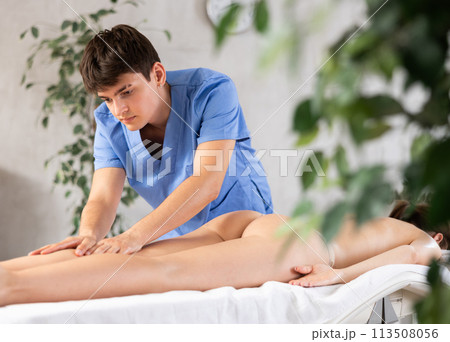 Male physiotherapist in medical suit massages legs thighs and calf muscles of girl client Male physiotherapist in medical suit massages legs thighs and calf muscles of girl client 113508056