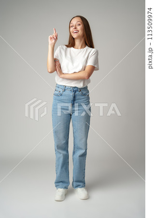 Portrait of pretty young woman in white T-shirt pointing up on gray background 113509974