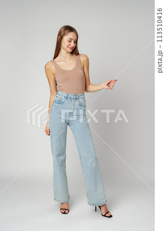 Young Woman Wearing High Rise Jeans and Tank Top posing on gray background in studio Young Woman Wearing High Rise Jeans and Tank Top posing on gray background in studio 113510416