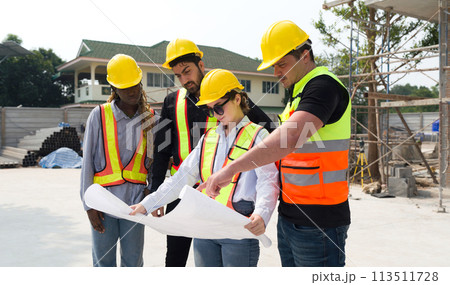 Group of construction workers gathered at a construction site reviewing some plan. Unfinished building, piles of construction material, and a partially constructed structure are in the background Group of construction workers gathered at a construction site reviewing some plan. Unfinished building, piles of construction material, and a partially constructed structure are in the background 113511728