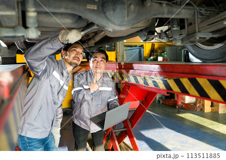 Two mechanic in gray uniform maintenance or inspection work on a vehicle that is raised on a lift while holding laptop computer. Diagnostic, referencing repair manual, or updating service record. Two mechanic in gray uniform maintenance or inspection work on a vehicle that is raised on a lift while holding laptop computer. Diagnostic, referencing repair manual, or updating service record. 113511885