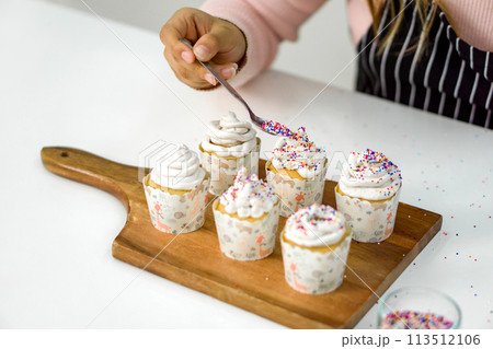 Closeup woman hand holding spoon with multicolor frosting, decorating cupcake on a wooden board, adding a colorful touch to the cupcakes' presentation. 113512106