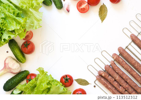 Healthy fresh vegetables for salad and raw sausages for barbecue. Raw green lettuce, greens, tomato over white countertop. Top view. Copy space. Food concept. Healthy fresh vegetables for salad and raw sausages for barbecue. Raw green lettuce, greens, tomato over white countertop. Top view. Copy space. Food concept. 113512312