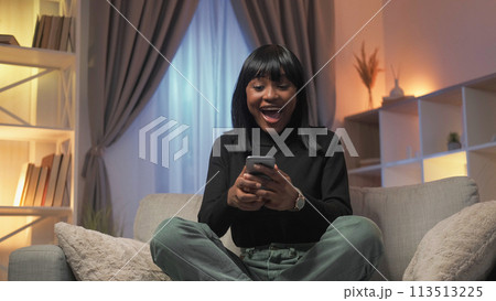 Online victory. Winner woman. Lucky giveaway. Excited surprised happy female looking smartphone celebrating success sitting home room interior. 113513225