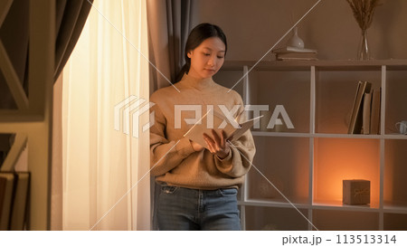 Interesting book. Curious woman. Weekend rest. Relaxed female enjoying reading staying near window in light room interior. 113513314