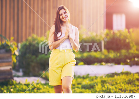Knee length woman portrait wearing yellow bermuda shorts, young caucasian female stands against blurred rural background on summer evening before sunset. Knee length woman portrait wearing yellow bermuda shorts, young caucasian female stands against blurred rural background on summer evening before sunset. 113516907
