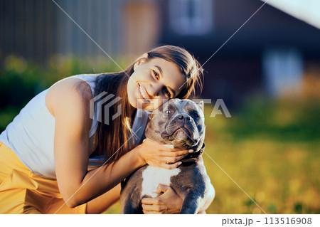 Dog is forced to look at camera with its head forcibly lifted, smiling portrait of young European woman with an American Bully in countryside at end of summer day. Dog is forced to look at camera with its head forcibly lifted, smiling portrait of young European woman with an American Bully in countryside at end of summer day. 113516908