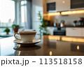 A close-up shot of a white coffee cup resting on a shiny kitchen counter with a blurred contemporary kitchen setting behind it, suggesting an early morning atmosphere. 113518158