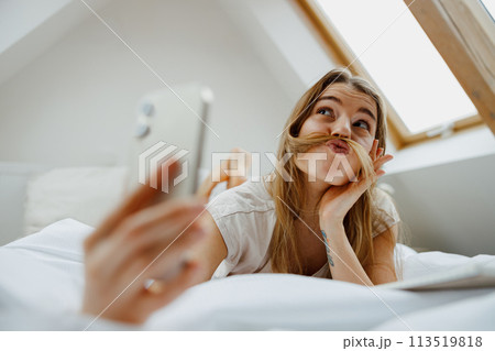 Smiling woman on bed, taking selfie with phone in comfort 113519818