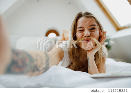 A woman is sitting comfortably on the bed, smiling and taking a selfie with her phone A woman is sitting comfortably on the bed, smiling and taking a selfie with her phone 113519821