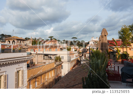 Scenic rooftop terrace view from luxury hotel in Rome. Outdoor furniture, green plants, sunny cityscape with warm toned buildings and Italian stone pines. Serene atmosphere. 113525055
