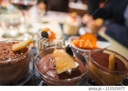 Close up view of assorted desserts in glass cups, featuring mousse and cream based dishes with various toppings. Background hints at dining setting, possibly a restaurant or formal experience. 113525077