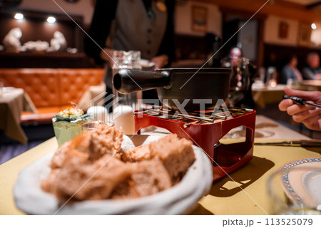 Dining scene with traditional fondue setup bread cubes on plate, black fondue pot on red stand with burner, interactive dining. Cozy restaurant ambiance, no identified location. Dining scene with traditional fondue setup bread cubes on plate, black fondue pot on red stand with burner, interactive dining. Cozy restaurant ambiance, no identified location. 113525079