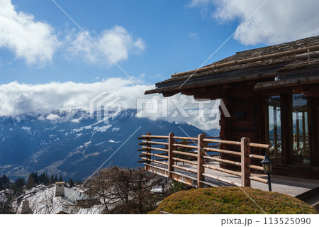 Rustic wooden chalet with balcony overlooking snowy peaks in mountainous region. Traditional lantern light, serene atmosphere. Ideal mountain retreat. 113525090
