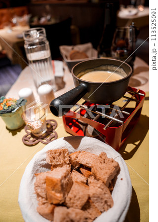 Cozy dining setting with fondue meal basket of bread cubes, whole grain variety, fondue pot on red stand with creamy mixture, wine glass, salt shakers, warm interior, intimate atmosphere. Cozy dining setting with fondue meal basket of bread cubes, whole grain variety, fondue pot on red stand with creamy mixture, wine glass, salt shakers, warm interior, intimate atmosphere. 113525091