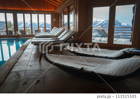 Indoor pool area with wooden interior, high ceiling, snowy mountain view through large windows, unique lounge chairs, sun loungers, luxurious mountain resort ambiance. Indoor pool area with wooden interior, high ceiling, snowy mountain view through large windows, unique lounge chairs, sun loungers, luxurious mountain resort ambiance. 113525105