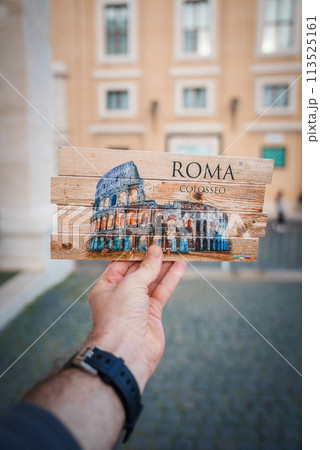 Hand holding up postcard with Colosseum, Rome, Italy image on rustic wooden background. ROMA and COLOSSEO printed. Urban setting, possibly in Rome. Watch suggests daytime. 113525161