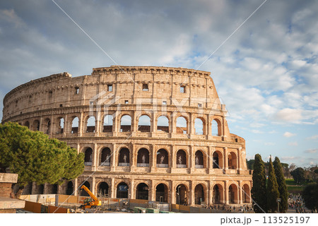 Colosseum in Rome, Italy. Iconic ancient amphitheater under soft, cloudy daylight. Arch shaped ruins with construction work, tourists, and trees nearby. Colosseum in Rome, Italy. Iconic ancient amphitheater under soft, cloudy daylight. Arch shaped ruins with construction work, tourists, and trees nearby. 113525197