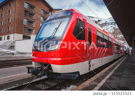 Modern red train at platform, likely part of Glacier Express in Switzerland. Sleek design with large front window for wide view. Snowy mountainous backdrop near Zermatt. Modern red train at platform, likely part of Glacier Express in Switzerland. Sleek design with large front window for wide view. Snowy mountainous backdrop near Zermatt. 113525322