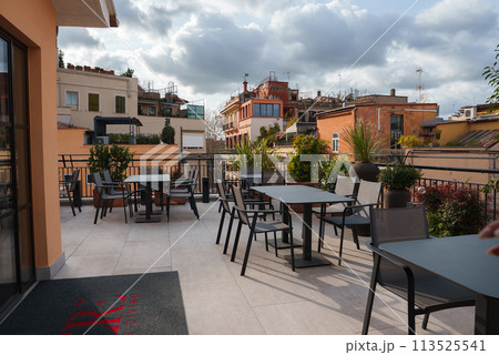 Luxury hotel rooftop terrace in Rome with dark colored furniture neatly arranged on light tile flooring. Traditional Roman architecture in the background. 113525541