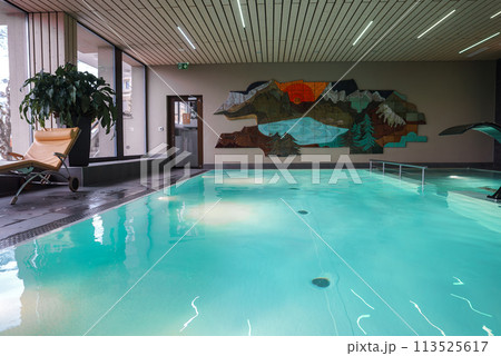 Indoor swimming pool area featuring calm turquoise waters, deck chair, potted plant, large windows, abstract wall art, wooden ceiling slats, tranquil ambiance. Indoor swimming pool area featuring calm turquoise waters, deck chair, potted plant, large windows, abstract wall art, wooden ceiling slats, tranquil ambiance. 113525617