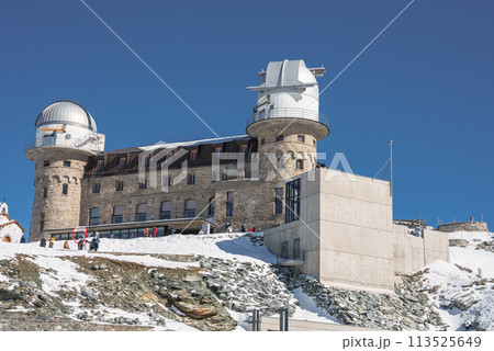 Grand stone building with traditional and modern elements on snowy landscape in Zermatt ski resort, Switzerland. Tower, observatory or weather station, people in winter clothing. Grand stone building with traditional and modern elements on snowy landscape in Zermatt ski resort, Switzerland. Tower, observatory or weather station, people in winter clothing. 113525649