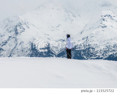 In Verbier, Switzerland, a lone figure in winter attire stands in snow, looking at rugged mountains under a cloudy sky, showcasing the serene alpine vastness. In Verbier, Switzerland, a lone figure in winter attire stands in snow, looking at rugged mountains under a cloudy sky, showcasing the serene alpine vastness. 113525722